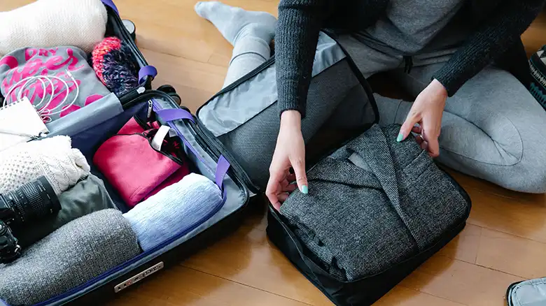 Hands beside an open suitcase containing folded clothing and sunglasses secured by packing straps