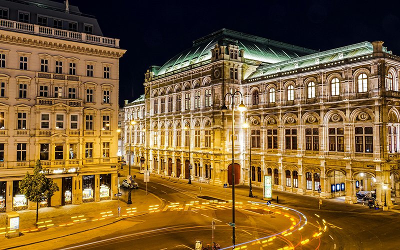 a street with lights on it and a large building with arched windows with Vienna State Opera in the background